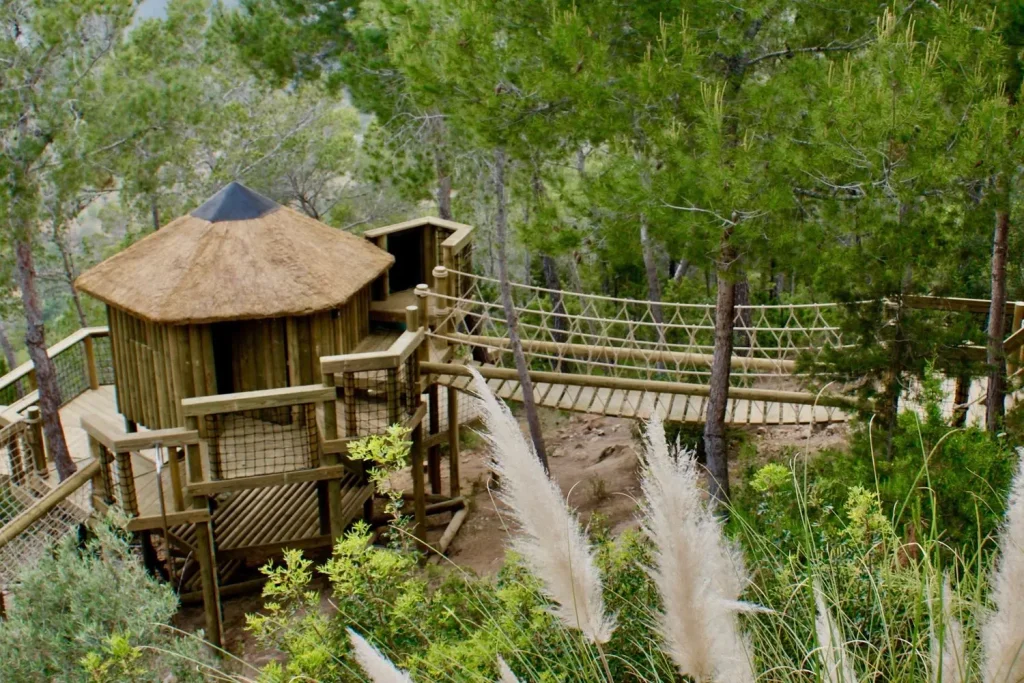Thatched roof garden Treehouse for the whole family with a fixed-beam Rope Bridge entrance leading to playful deck platforms wrapping a sleep-over Treehouse, all within a mediterranean forest and woodland setting on a gradient.