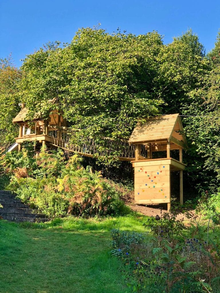 Two Treehouse play tower platforms set into a garden gradient amongst trees with Rope Bridge, Slide and Climbing Wall