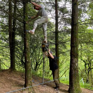 Two Rope Bridge installers using ground anchor install kit, one holding jack hammer with the other standing on top, in pine woodland, Northern Ireland.