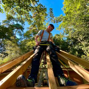 Treehouse builder person installing a Treehouse roof, sat high up on the roof joists and beams with fixing equipment and tools.