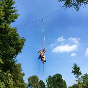 Tree Climbing cable installer suspended mid-air in woodland setting from a galvanised steel, cable between trees, wearing a harness and safety equipment.