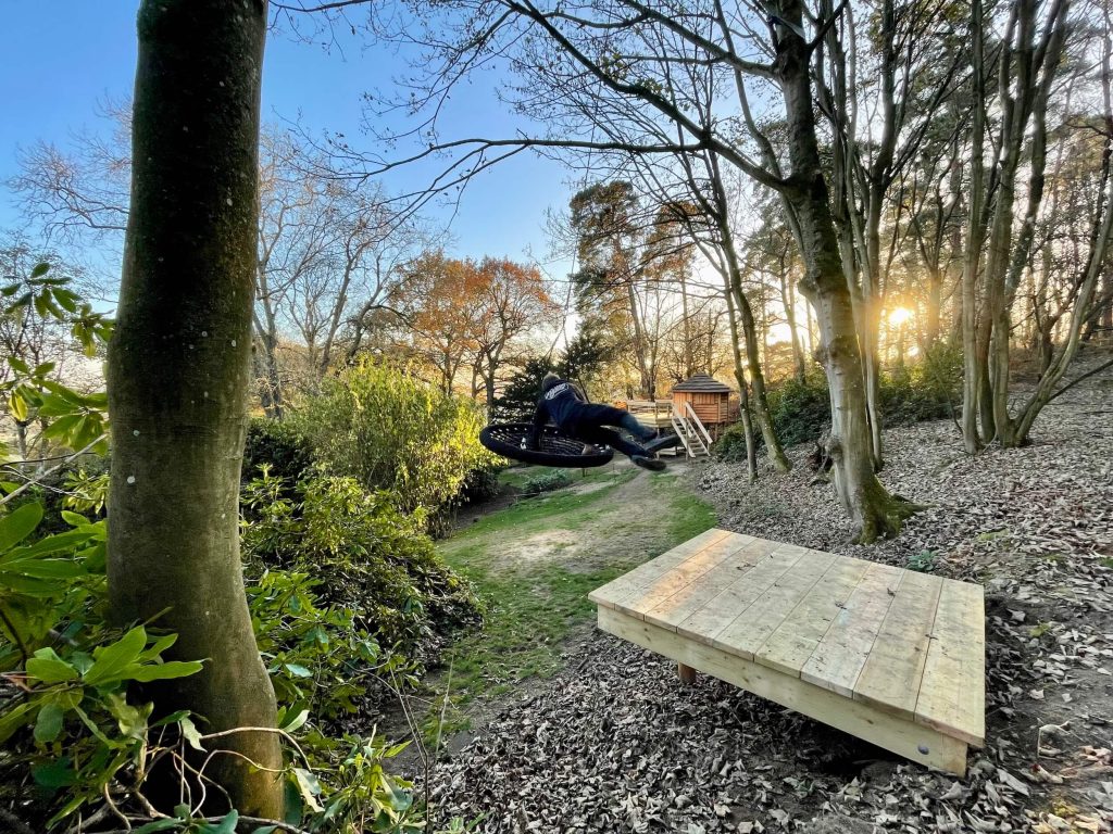 Woodland scene in Surrey, with a Treehouse in the distance and a person in black leaping from woodland Deck Platform on to a suspended Nest Swing.
