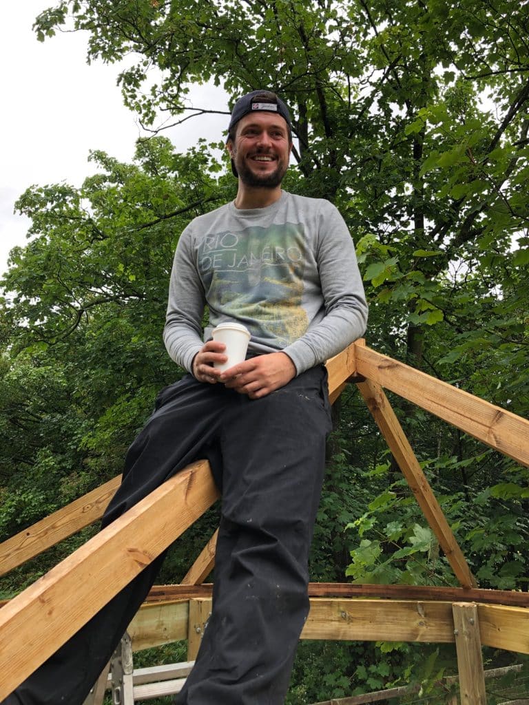 A man sitting on the top of a roof frame for a garden treehouse holding a cup of coffee and smiling at the camera.