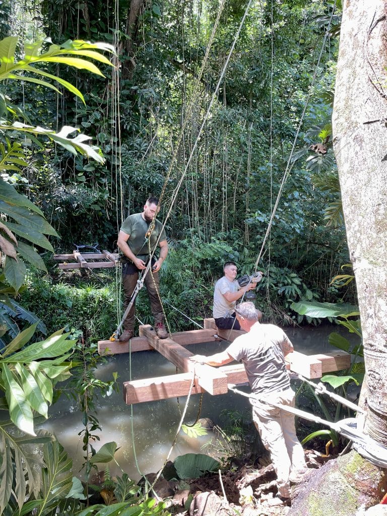 3 members of the Treehouse Life team are fixing suspension ropes to hoist large beams over a river in the Hawaiian jungle. The beams will form the main structure of a Rope Bridge in a luxury commercial resort.
